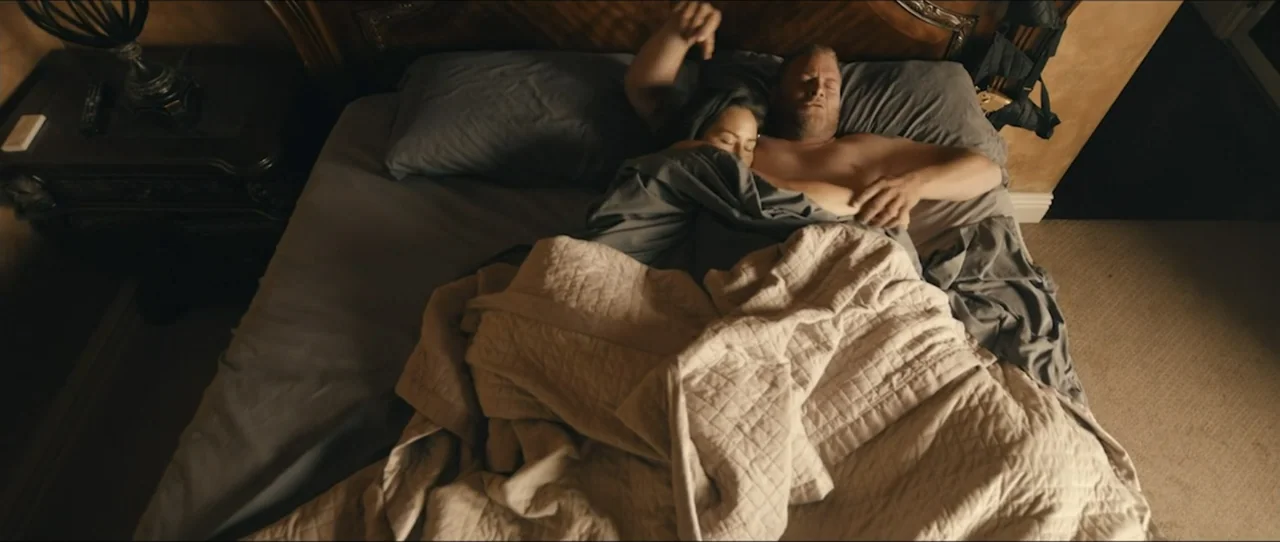 American Sicario (2021) movie still: wide — A dark bedroom with a large bed featuring grey pillows and a quilted blanket. A man and woman sleep side…