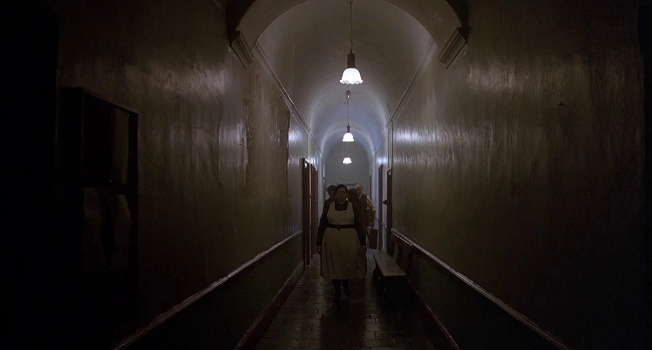 Cracks (2009) movie still: wide — A long, vaulted hospital corridor features painted walls and a row of pendant lamps. A woman in an apron walks…