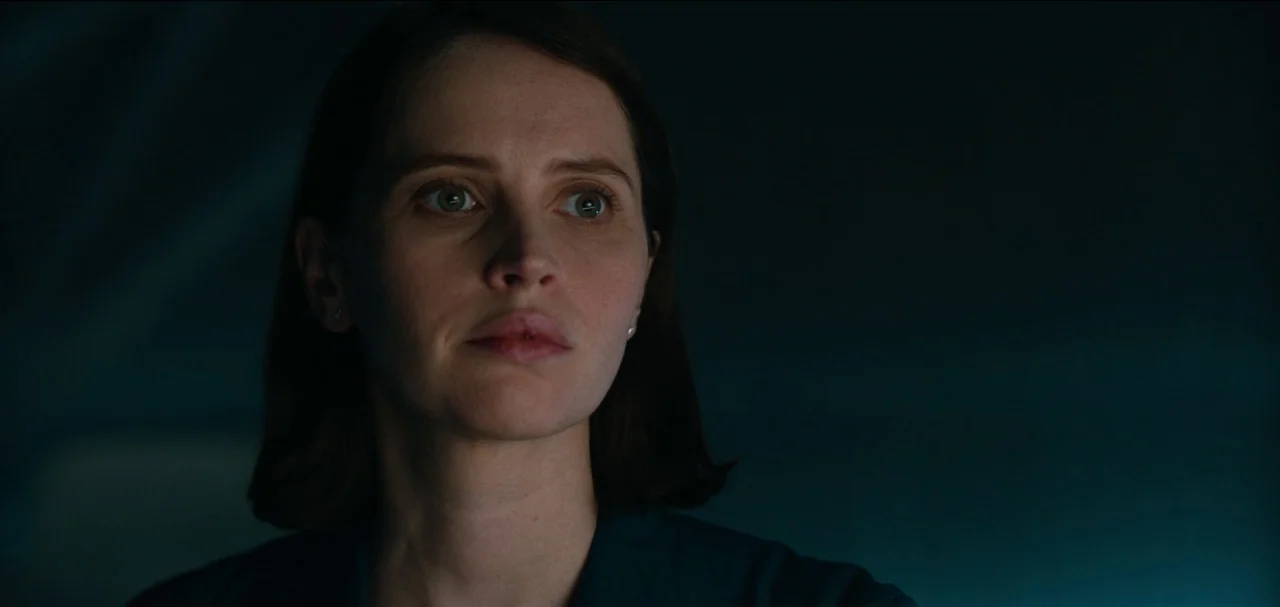 The Midnight Sky (2020) movie still: closeup — A dimly lit interior space features a dark, unadorned background. A woman looks off-camera while…