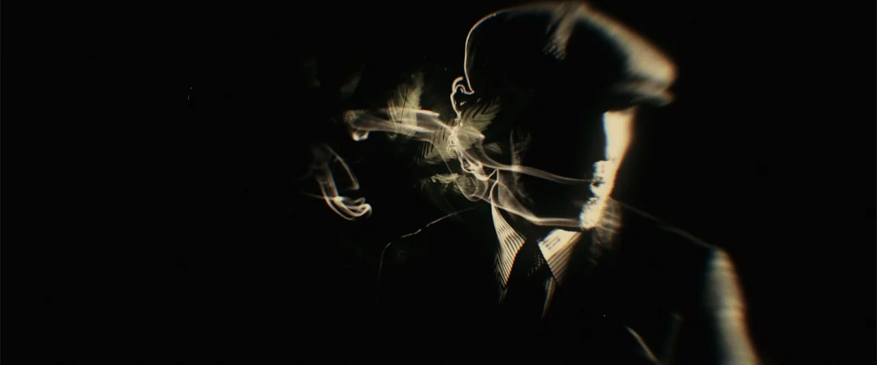 The Gentlemen (2020) movie still: closeup — A dark space features a man wearing a cap partially illuminated by light. Trails of smoke drift across…