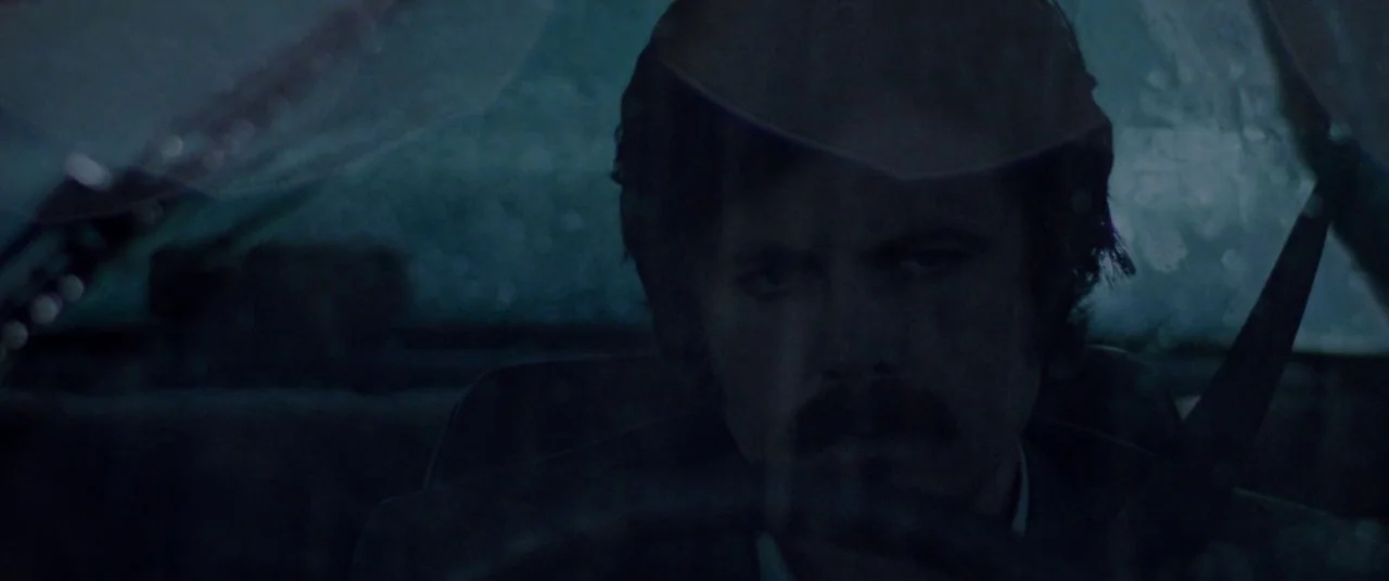 The Old Man & The Gun (2018) movie still: closeup — A man sits behind the steering wheel of a car at night. Rain streaks the windshield while he…