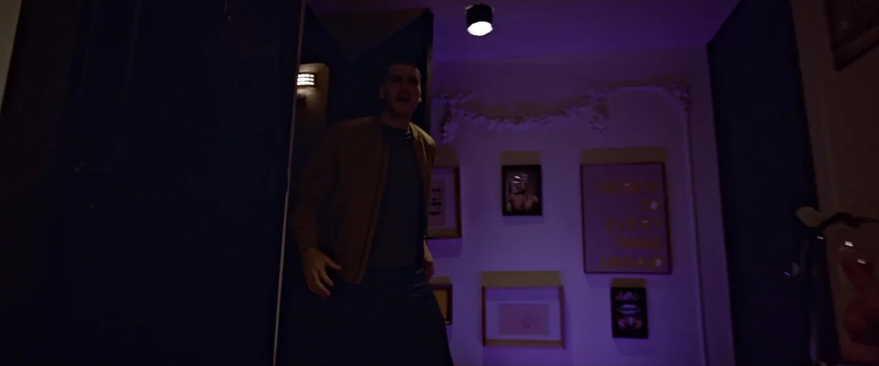 Like or Die (2023) movie still: medium — A residential hallway features white walls adorned with framed pictures and a motivational sign. A young…