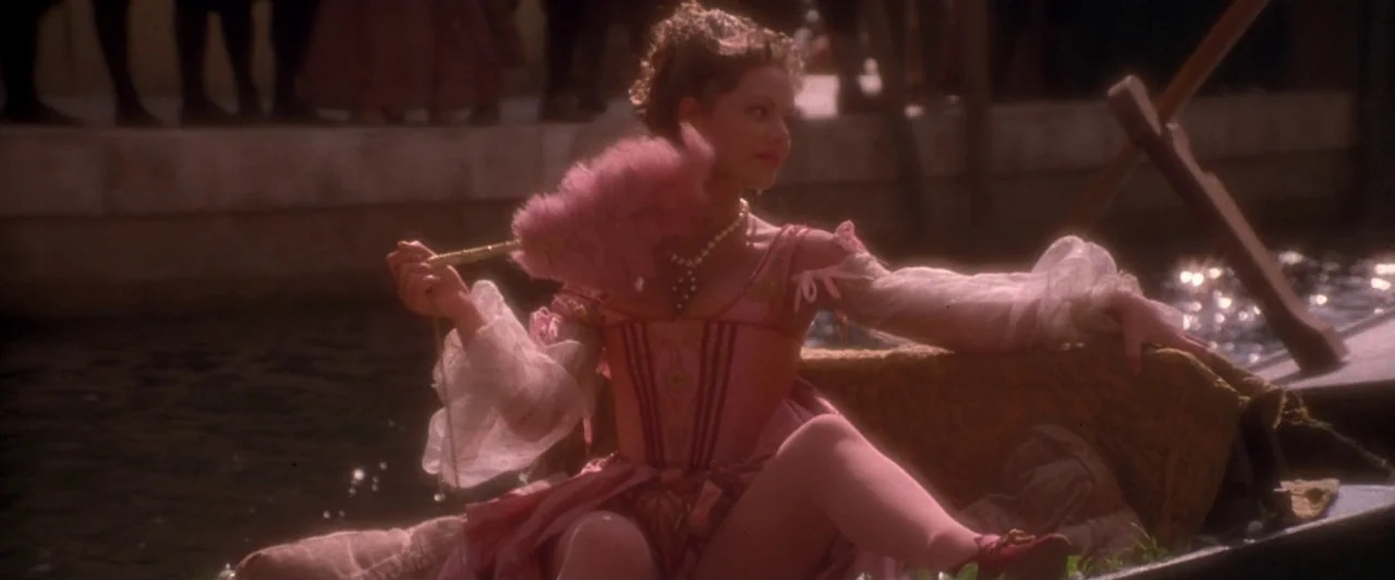 Dangerous Beauty (1998) movie still: medium — A woman sits inside a gondola floating on a sunlit Venetian canal. She holds a small pink feathered…