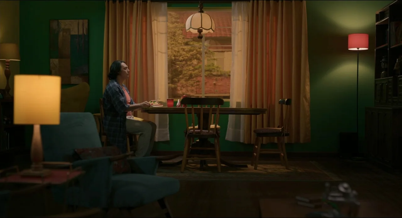 The Beauty and the Moody (2022) movie still: medium — A green-walled dining area contains a wooden table, chairs, and multiple lit floor lamps. A…