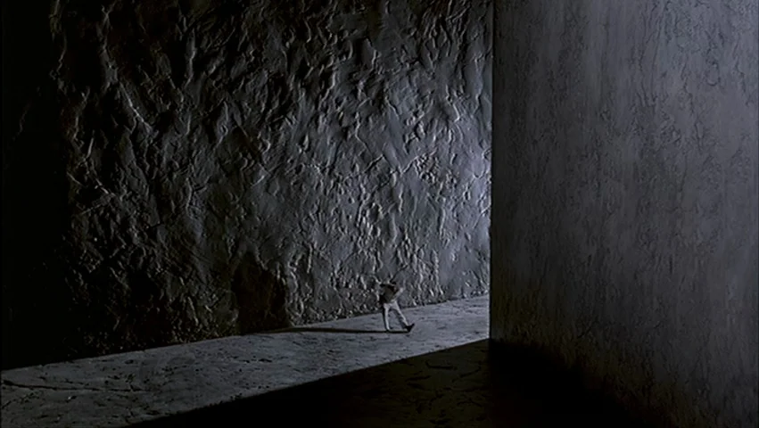 The Big Lebowski (1998) movie still: wide — A dark corridor features highly textured, uneven walls and a flat concrete floor. A single person walks…