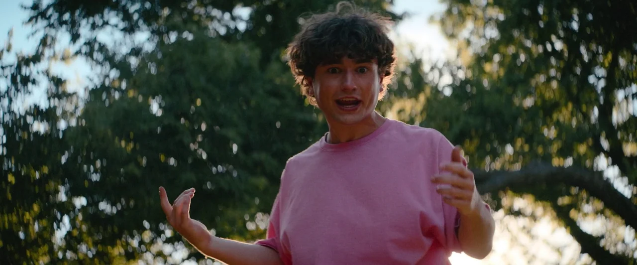 Snack Shack (2024) movie still: medium — A man stands outdoors in front of out-of-focus trees during the late afternoon. He is actively talking with…