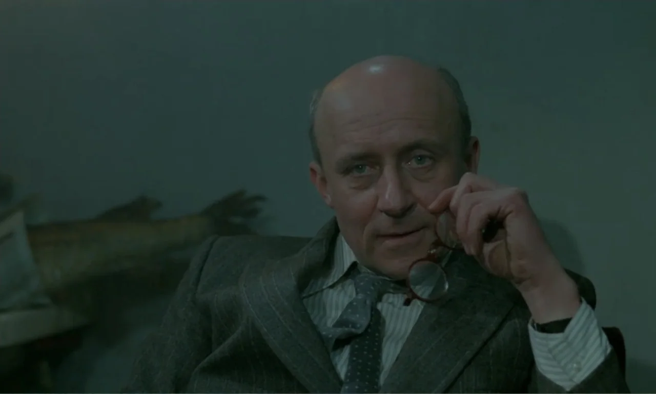 Ga-ga: Glory to the Heroes (1986) movie still: closeup — A dimly lit office features a textured wall and a large fish mount visible. A bald…