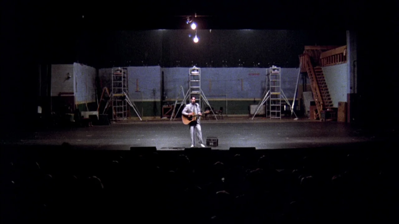 Stop Making Sense (1984) movie still: wide — An empty theater stage with exposed scaffolding and a wooden staircase in the back. A lone musician…