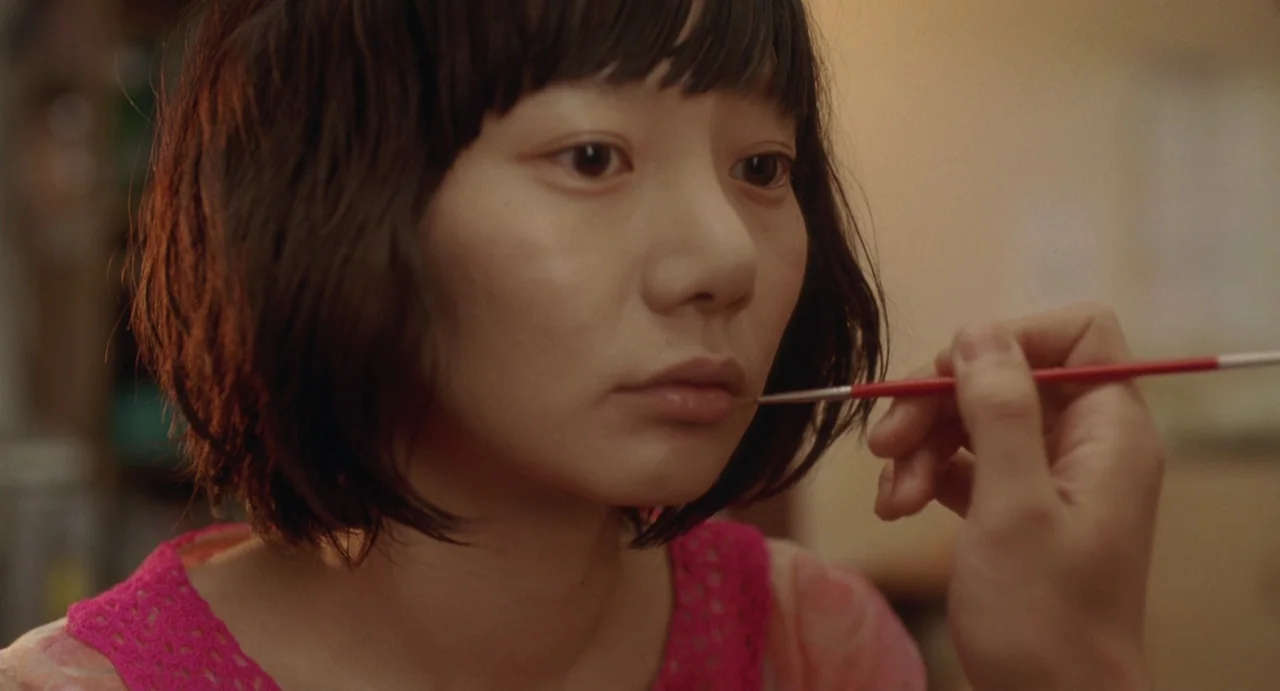 Air Doll (2009) movie still: closeup — An interior space features a woman receiving makeup application on her lips. A hand holds a small red-handled…