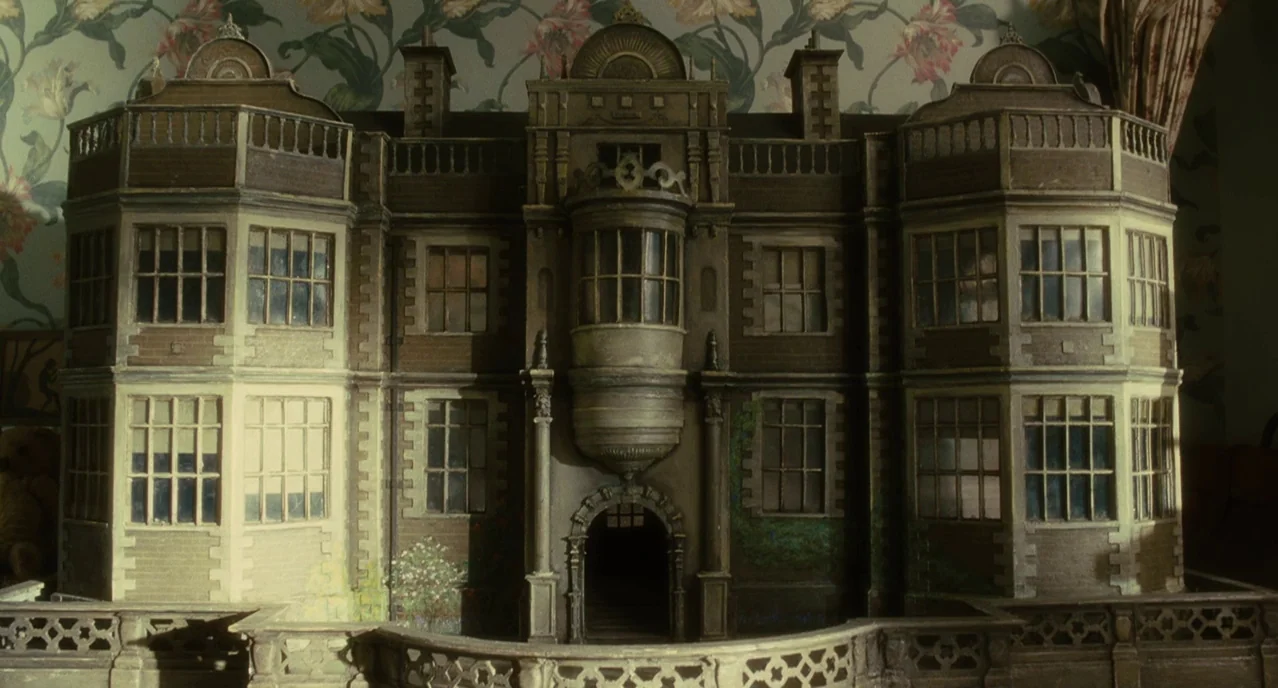 Atonement (2007) movie still: medium — An ornate wooden dollhouse rests against a wall decorated with vintage floral patterned wallpaper. The…