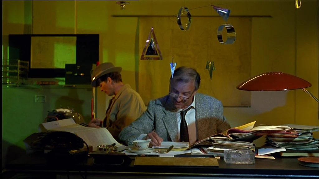 Lola (1981) movie still: medium — An office filled with paperwork under a harsh, green-tinted lighting setup. Two men work at a large desk cluttered…