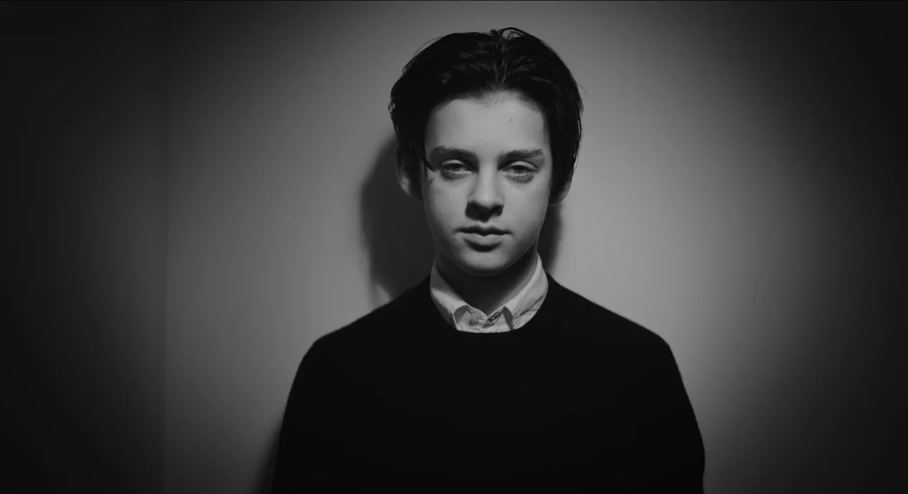 One More Time With Feeling movie still: medium — A person stands centered against a neutral wall with heavy side lighting present. The subject looks…