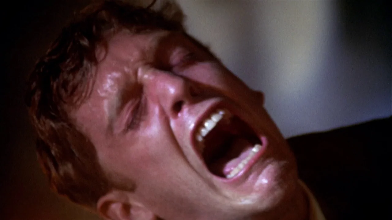 The Day of the Locust (1975) movie still: closeup — A close-up shot focuses on a man's face in a dark, indistinct environment. He is shouting with…