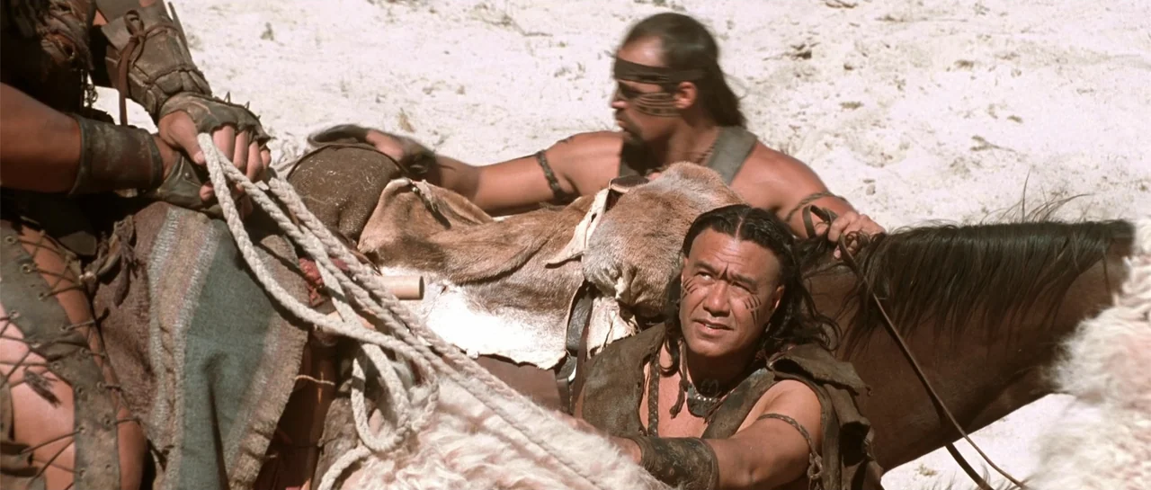 The Scorpion King (2002) movie still: medium — Two men on horseback stand in a bright, sandy, featureless desert environment. The men wear primitive…