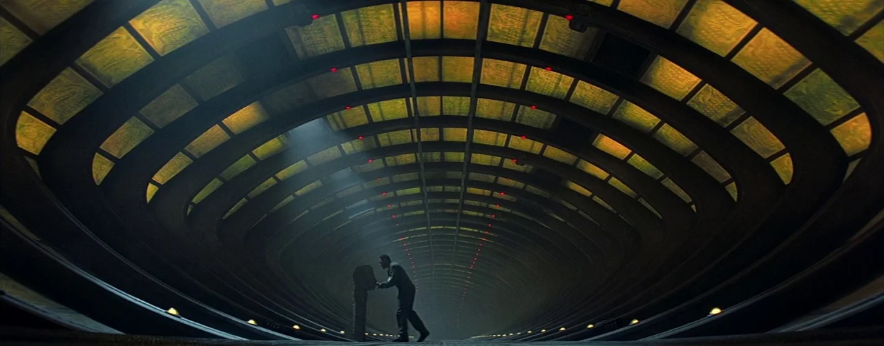 Event Horizon (1997) movie still: wide — A long, curved, elliptical corridor features glowing amber ceiling panels and dark metal surfaces. A lone…