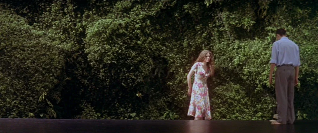 Talk To Her (2002) movie still: fullbody — A dense, overgrown green hedge serves as a backdrop for two people outdoors. The man stands with his back…
