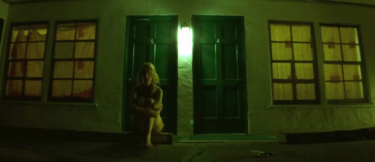 Tangerine (2015) movie still: wide — A dimly lit motel walkway at night features two numbered doors and two windows. A woman sits on the ground in…