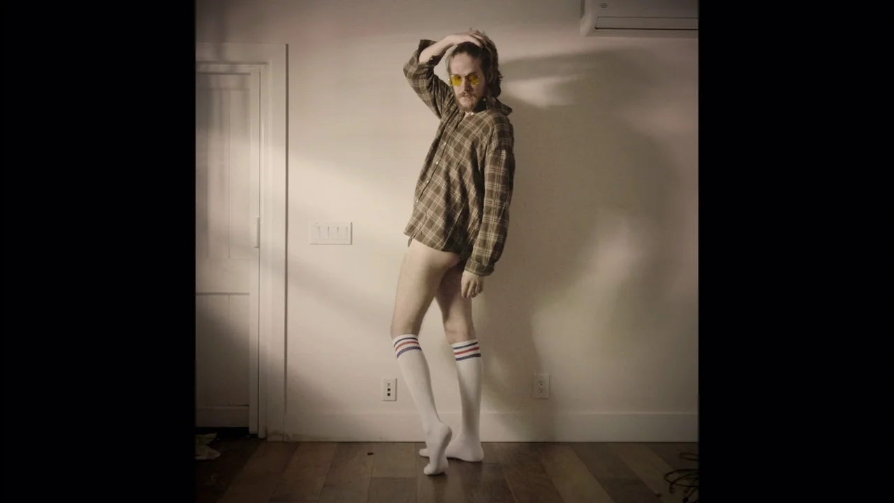 Bo Burnham: Inside (2021) movie still: fullbody — A spare interior space features a plain white wall and wood flooring. A man stands posing barefoot…
