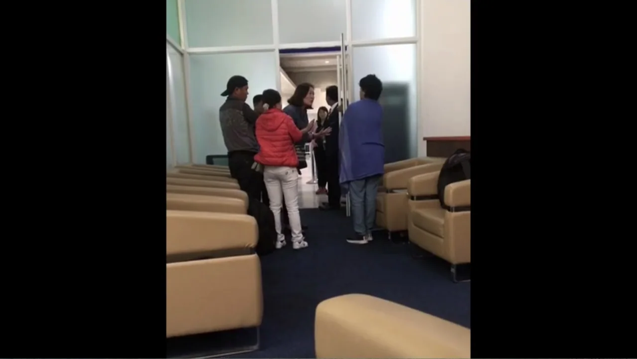 Escape From Saudi (2019) movie still: fullbody — A modern building waiting area with tan armchairs and white framed glass doors. Five people stand…