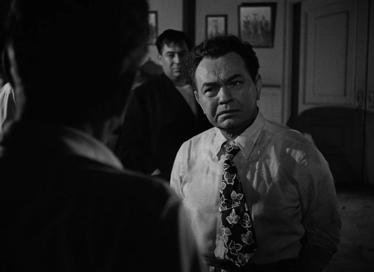 Key Largo (1948) movie still: medium — A dimly lit room with framed pictures on the wall and a door frame. A man in a patterned tie looks towards…