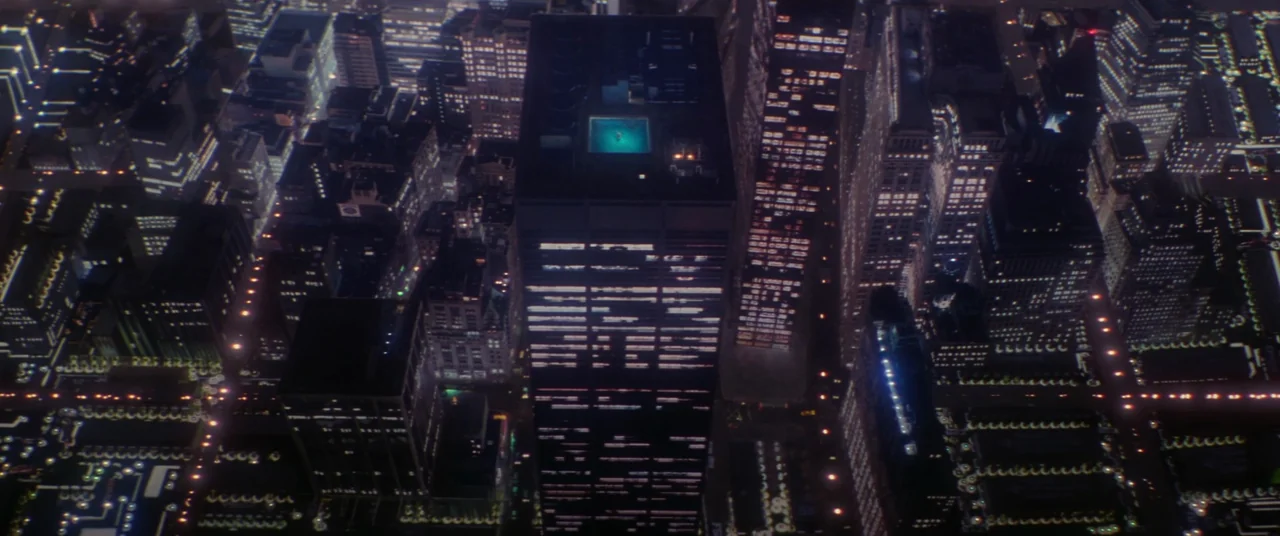 Hackers (1995) movie still: establishing — An aerial view looks straight down at a dark metropolis grid at night. A brightly lit cyan rooftop…