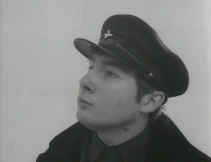 Les Carabiniers (1963) movie still: closeup — A plain white background occupies the space behind a young man. The man wears a peaked military cap…