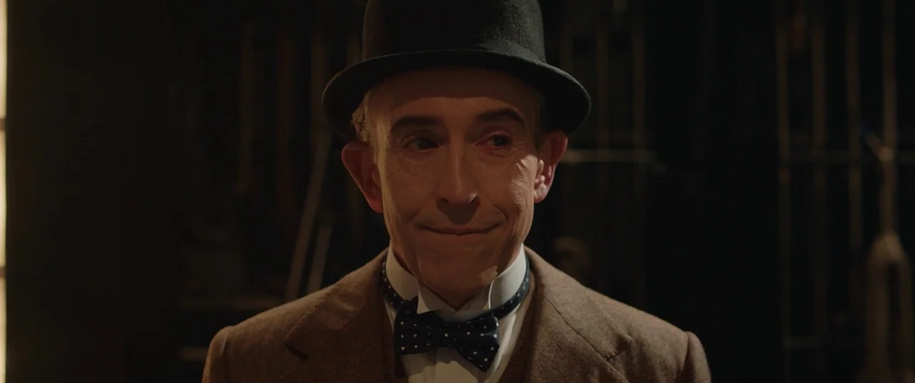 Stan & Ollie (2018) movie still: closeup — A man wearing a bowler hat stands in a dimly lit, dark space. The subject stares ahead with a closed,…