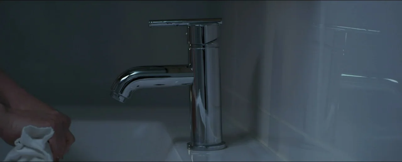 Hostages (2017) movie still: closeup — A modern bathroom features a white basin and a polished chrome faucet. A person's hand grips a white cloth…