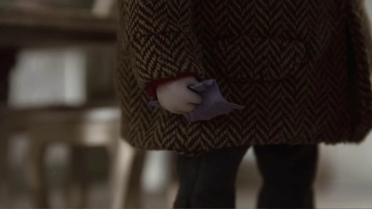 Bestia (2017) movie still: closeup — The interior shows a blurred background with furniture shapes. A small articulated hand holds a folded purple…