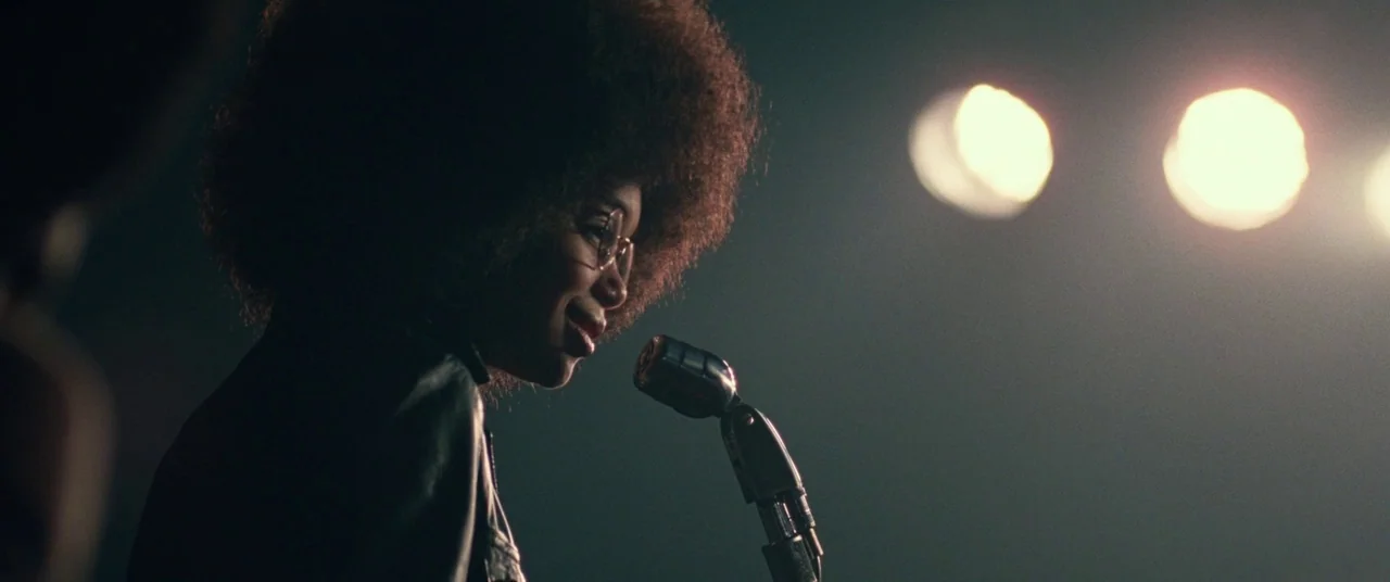 BlacKkKlansman (2018) movie still: medium — A dark concert stage lit by bright circular spotlights directed from the side. A young woman with an…
