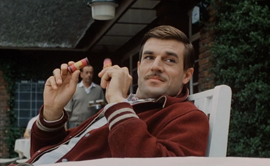 Soldier of Orange (1977) movie still: medium — A man sits on a patio outside a brick building with a thatched roof. He holds two small cylindrical…