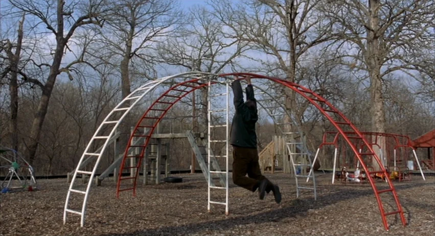 About Schmidt movie still: wide — An outdoor playground features a tall arched metal ladder structure over wood-chipped ground. A man hangs from the…