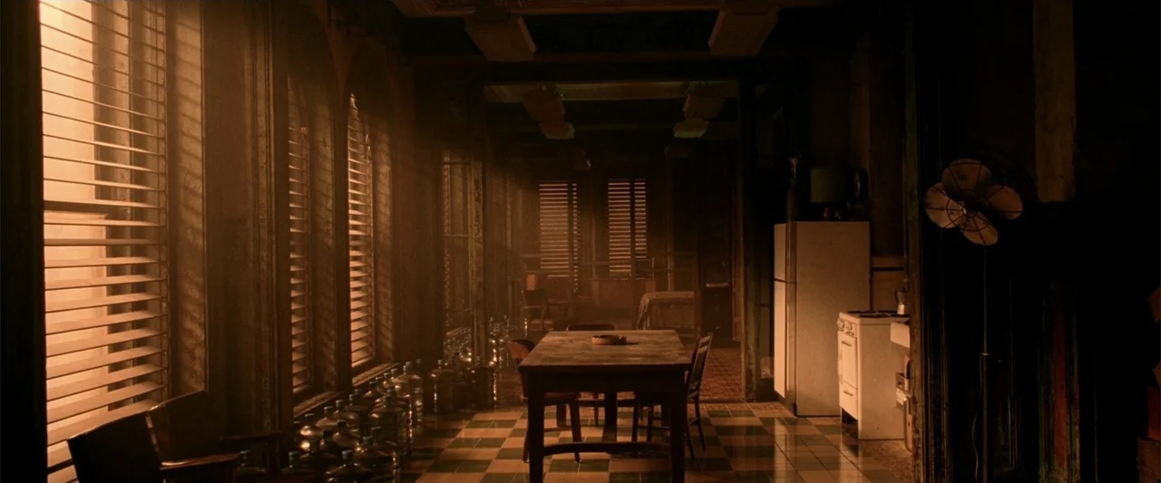 Constantine (2005) movie still: wide — A dimly lit room features a wooden table, a white refrigerator, and rows of water jugs. Blinds on the left…