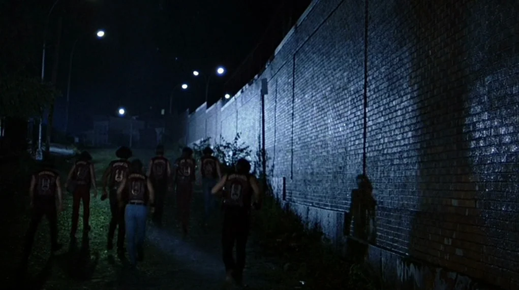 The Warriors (1979) movie still: wide — A group of men in matching vests run beside a long, looming brick wall. Streetlamps illuminate the path…