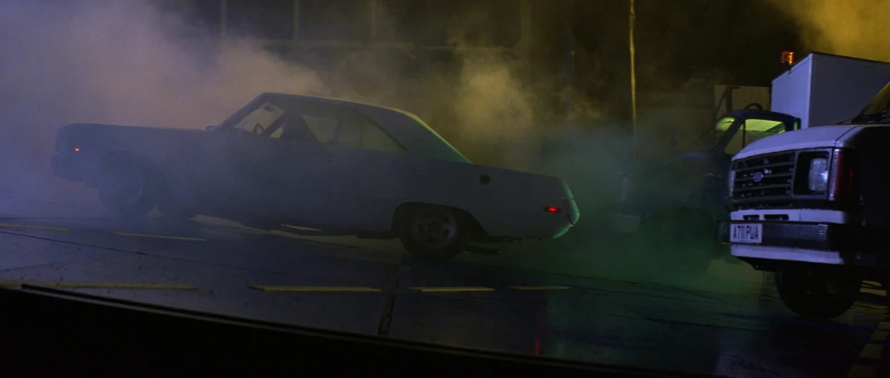 The Cook, The Thief, His Wife & Her Lover (1989) movie still: wide — A vintage coupe drives through thick, swirling smoke in a dark paved area. A…