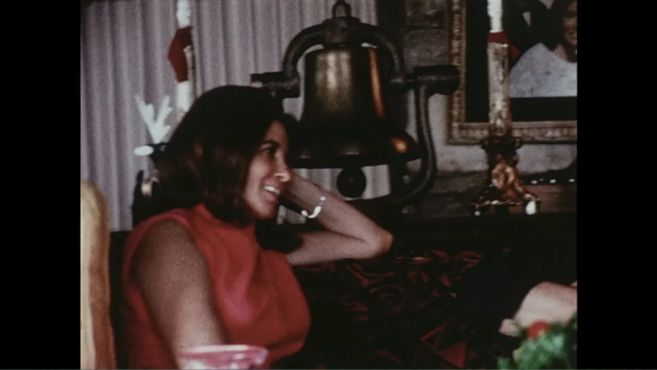 The Winding Stream (2014) movie still: medium — A dimly lit domestic interior features a decorative metal bell placed on a shelf. A woman with…