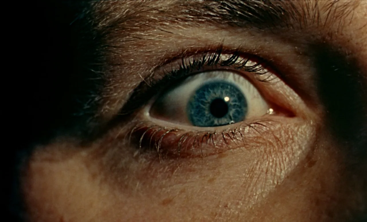Peeping Tom (1960) movie still: closeup — A close-up shot focuses on a single human eye against a dark background. The bright blue iris is sharply…