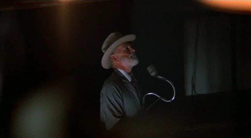 The Coca-Cola Kid (1985) movie still: medium — An older man wearing a hat stands at a podium in a dark room. He addresses an audience while facing a…