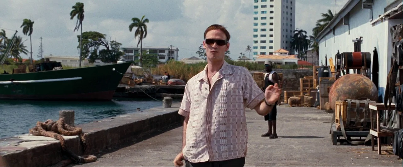 Quantum of Solace (2008) movie still: fullbody — A sunlit industrial wharf beside a harbor features a large cargo ship moored nearby. A thin man…