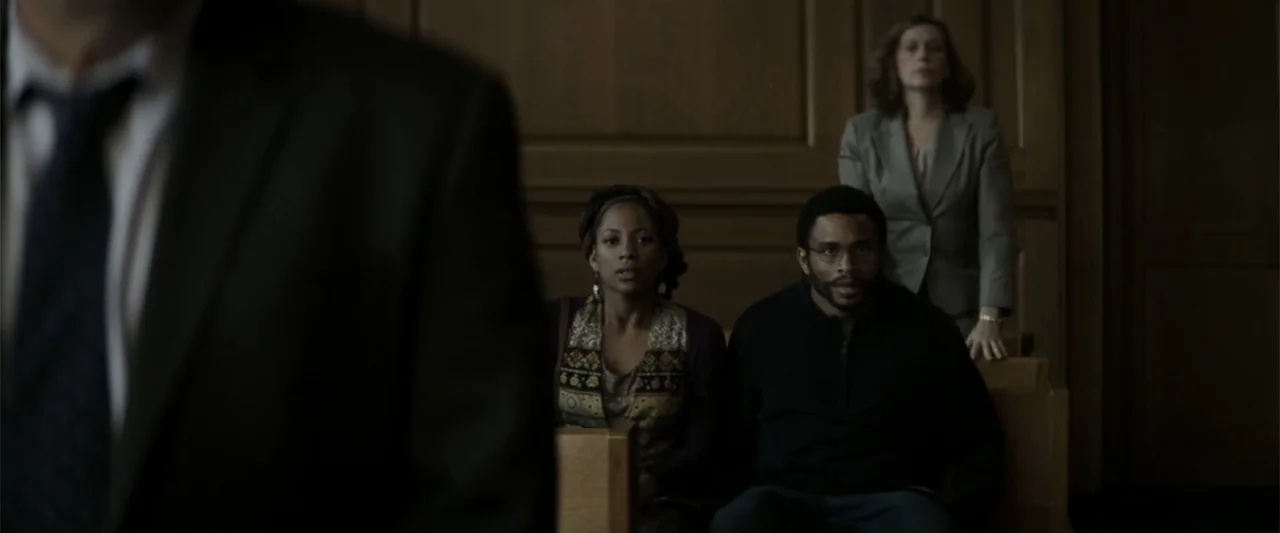 Crown Heights (2017) movie still: medium — A dark wooden courtroom contains rows of benches where people are seated. Three individuals sit facing…