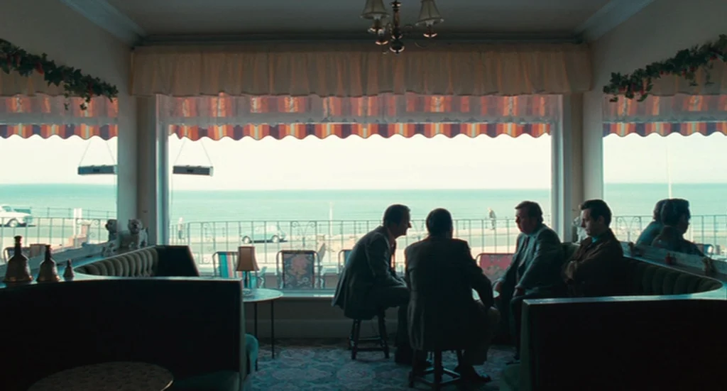 The Damned United (2009) movie still: wide — A seaside dining area features curved green velvet booths and a wide window view. Four men are seated…
