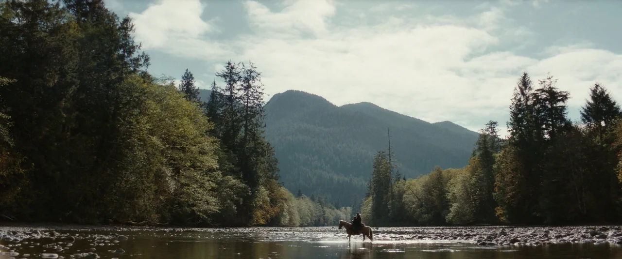 The Dead Don’t Hurt (2023) movie still: wide — A wide shot shows a person on horseback crossing a shallow, rocky river. Dense evergreen forests line…