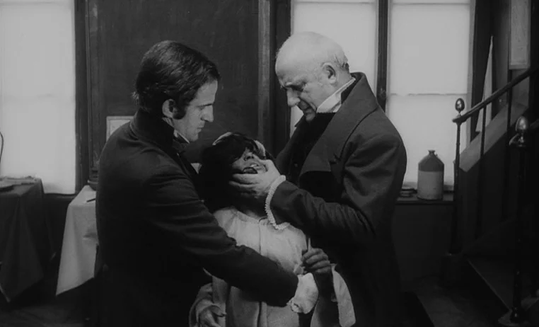 The Wild Child (1970) movie still: medium — A dimly lit room with plain walls, large window panes, and a wooden staircase. Two men in period coats…