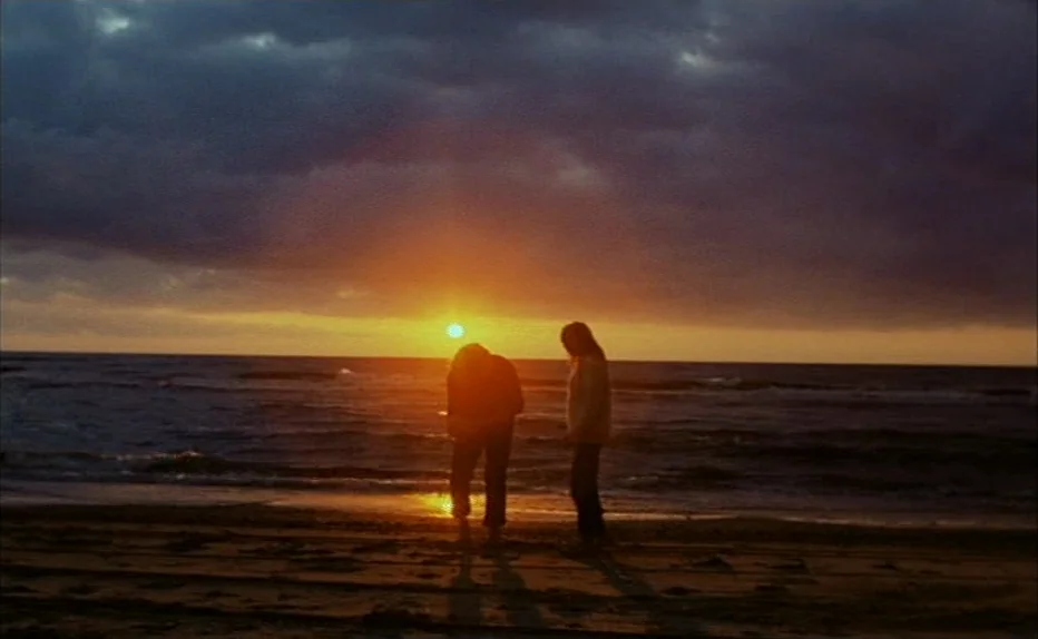 Turkish Delight (1973) movie still: wide — A wide beach shore is visible at golden hour under a heavy, dark cloud layer. Two figures stand close to…