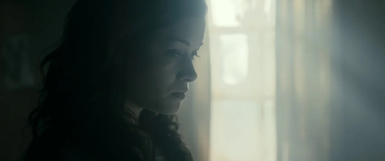 Evil Dead (2013) movie still: closeup — A dimly lit room features a woman positioned near a bright, light-filled window. A soft haze permeates the…