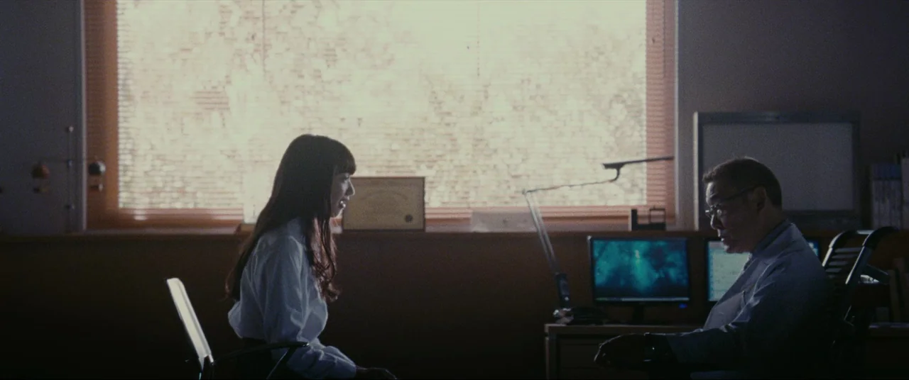The World of Kanako (2014) movie still: medium — A clinical consultation room features a window with horizontal blinds and two computer monitors. A…
