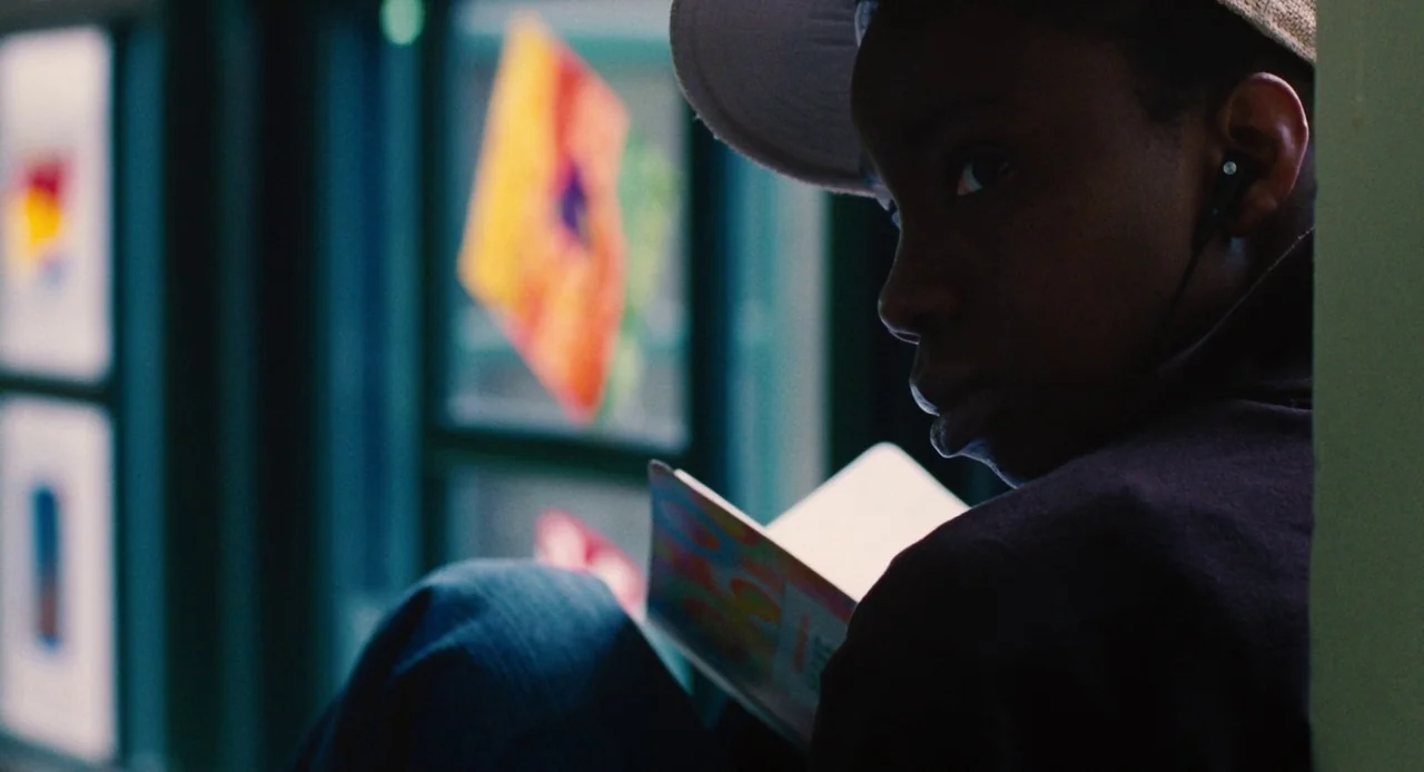 Pariah (2011) movie still: closeup — A person sits near a window inside a space decorated with colorful paper art. The individual holds an open book…