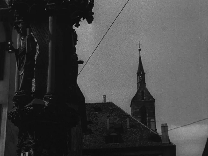 Thirst (1949) movie still: wide — A European city square features a dark, ornate stone monument in the immediate foreground. A traditional steeple…