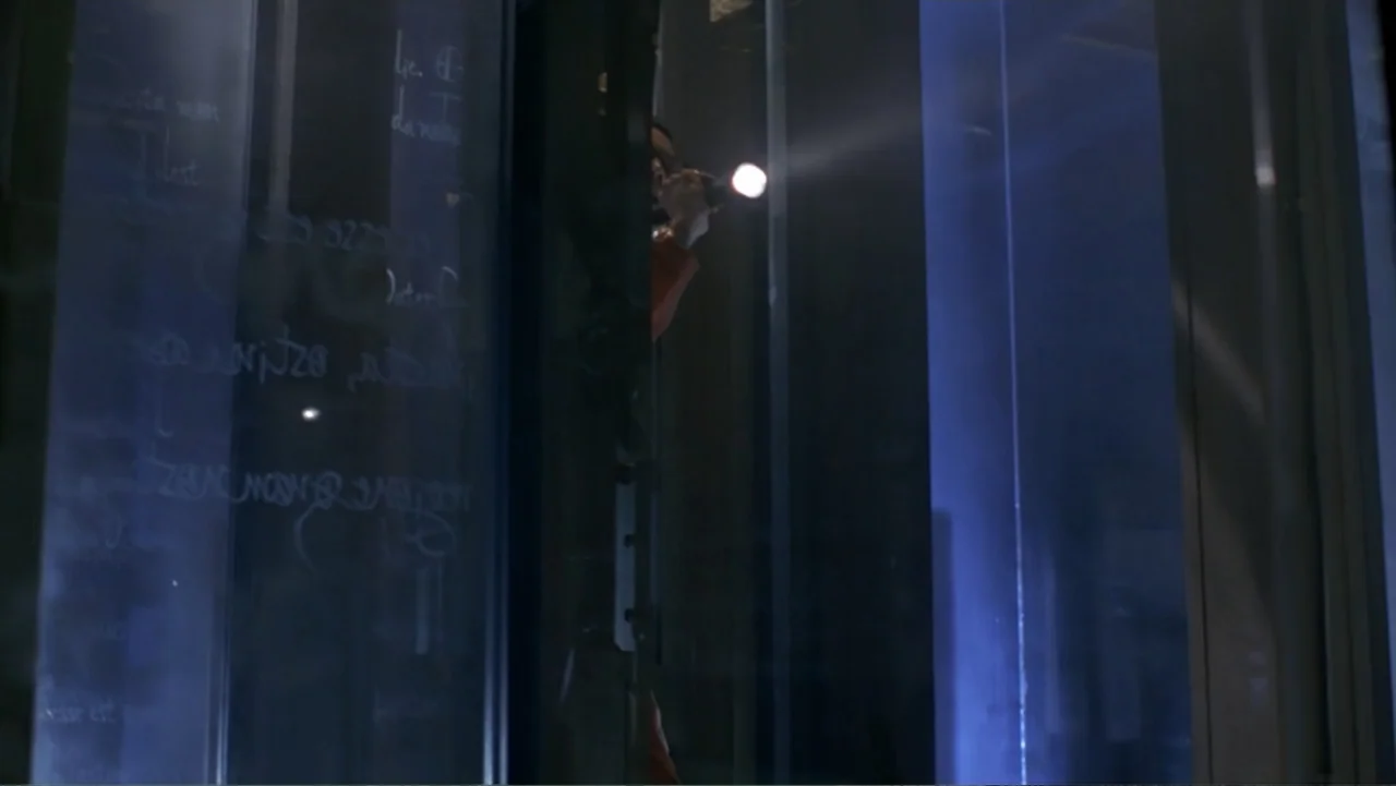 13 Ghosts (1960) movie still: medium — A dark space features a tall vertical pane of glass with faint white lettering. A person partially obscured…