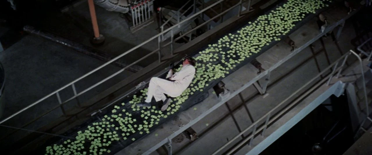 Soylent Green (1973) movie still: wide — A high-angle view of an industrial facility with a long moving conveyor belt system. A man in white…